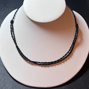 May include: A black cord necklace featuring a double-strand design made from thin, twisted material. The necklace includes two small, adjustable sliding knots on either side, allowing for length customization to fit the wearer comfortably.
