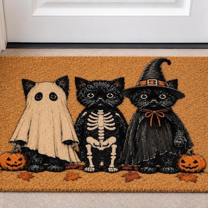 May include: A rectangular tan coir doormat featuring a printed illustration of three black kittens. The left kitten wears a ghost sheet, the center kitten has a skeleton print, and the right kitten wears a witch hat and cape with two pumpkins.