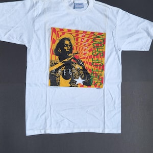 May include: A white short-sleeve t-shirt featuring a graphic print of a person wearing a hat and singing into a microphone. The background of the graphic is yellow and orange with a wavy pattern. The text reads GREGORY ISAACS STATE OF SHOCK.
