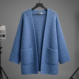 May include: A periwinkle blue open-front cardigan made of thick, textured crochet yarn. This long-sleeved garment features a relaxed fit, a straight hemline, and two large, square patch pockets on the front. It is displayed on a black hanger.