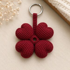 May include: A dark red crochet keyring featuring four stuffed heart-shaped elements arranged in a clover pattern. The hearts are connected to a central circular ring and attached to a silver-toned metal split ring at the top by a short crocheted strap.