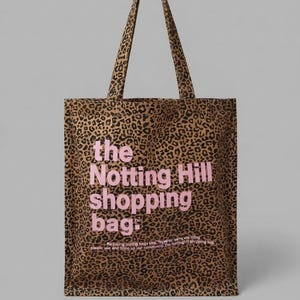 May include: A brown and black leopard print tote bag with two shoulder straps. The text "the Notting Hill shopping bag" is printed in large pink letters on the front, with smaller text below it regarding plastic use and community.