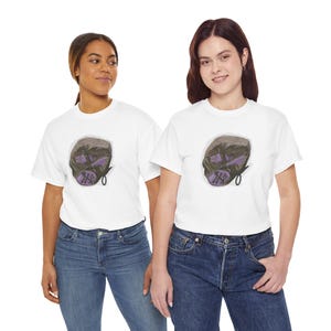 May include: A white short-sleeved t-shirt featuring a central graphic of a stylised, dark-toned mask with purple accents and a small black loop detail. The design is centred on the chest area of the garment, which is made of a plain white fabric.