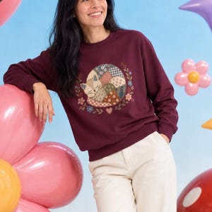 May include: A maroon long-sleeve sweatshirt featuring a graphic of a curled-up cat with a patchwork pattern, surrounded by small floral elements. The sweatshirt is paired with cream-coloured trousers and white trainers.