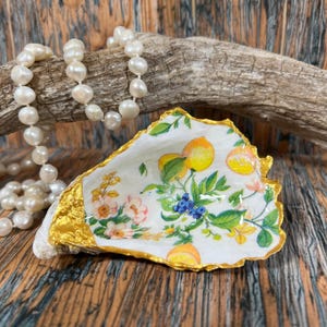 May include: A decorative oyster shell dish featuring a painted pattern of yellow lemons, green leaves, and pink flowers on a white interior with a metallic gold painted rim, accompanied by a strand of white freshwater pearls.