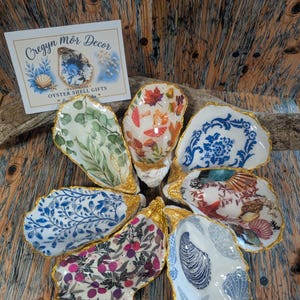 May include: A collection of seven decorative oyster shells with gold-painted edges, each featuring unique patterns including botanical leaves, autumn mushrooms, blue floral motifs, and seashells. A white card labelled Cregyn môr Decor is included.