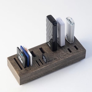 May include: A rectangular dark wood storage organiser for memory cards and flash drives. The surface features laser-engraved labels for SD, Micro SD, USB, and Type-C slots. Several memory cards and three USB flash drives in black and white are inserted into the designated slots on the wooden base.