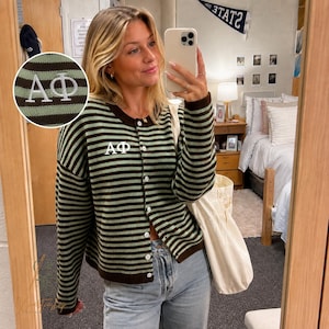May include: A long-sleeved button-down cardigan featuring horizontal stripes in dark brown and light green. The garment displays the white embroidered Greek letters Alpha Phi, or AΦ, on the upper left chest area. The cardigan has a round neckline.