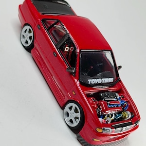 以下が含まれることがあります： エンジンルームが開いた詳細な赤いダイキャスト製モデルカー。シルバーの6本スポークホイールと黒いルーフが特徴です。フロントガラスには白い文字でTOYO TIRESと書かれています。インテリアには赤いハーネス付きのレーシングシートが装備されています。