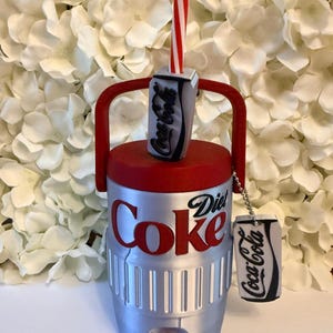 May include: A silver beverage cooler with a red lid and handle, featuring the text Diet Coke in red and black. A miniature soda can with a red and white striped straw sits on top, and a small Coca-Cola branded tag hangs from the handle by a chain.