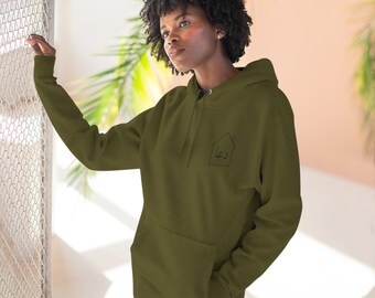 May include: An olive green hooded sweatshirt featuring a black line drawing of a house on the left chest. Inside the house outline, the Arabic word "دعا" is printed in black ink. The garment includes a front kangaroo pocket and a drawstring hood.