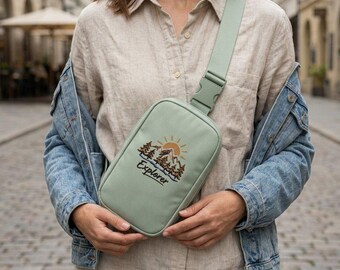 May include: A sage green crossbody sling bag featuring an embroidered graphic of a mountain range, pine trees, and a sun, with the word Explorer stitched below. The bag has a matching adjustable strap with a plastic buckle closure.