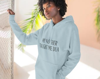 May include: A light blue hooded sweatshirt featuring the black text "MY MOTHER TAUGHT ME DUA" printed across the chest. The garment includes a front kangaroo pocket, long sleeves with ribbed cuffs, and a matching drawstring hood for a casual look.