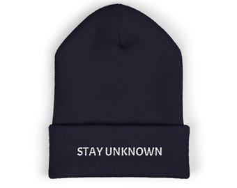 May include: A dark navy blue knit beanie with a folded cuff. The front of the cuff features the text STAY UNKNOWN embroidered in white capital letters. The hat has a classic cuffed design with a ribbed texture throughout the fabric.