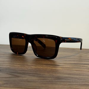 Könnte beinhalten: Prada-Sonnenbrille mit Schildpattmuster, dickem, rechteckigem Rahmen und dunkelbraunen Gläsern. Das Prada-Logo ist in der oberen Ecke des rechten Glases sowie auf dem äußeren Bügel des Rahmens deutlich zu erkennen.