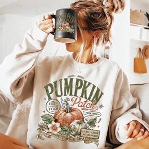 May include: A cream-colored crewneck sweatshirt featuring a large graphic print with the text PUMPKIN Patch, Fresh from the Patch, and Farm Fresh. The design includes illustrations of orange and white pumpkins, leaves, and flowers. A black mug with a matching pumpkin graphic is held up.