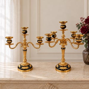 May include: Two gold-toned metal candelabras with dark blue accents. The left candelabra has three candle holders, while the right one has five. Both feature ornate, curved arms, a central stem, and a circular base decorated with gold floral patterns.