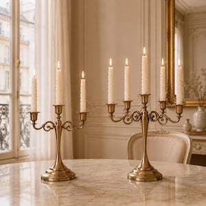 May include: Two brass candelabras with a polished finish. The left candelabra holds three lit white spiral taper candles, while the right candelabra holds five lit white spiral taper candles. Both feature curved arms and a circular base.