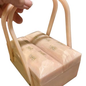 May include: A rectangular, light pink marbled handbag featuring two hinged lids that open from the center. The surface is adorned with small, gold-toned bee-shaped embellishments. It includes two matching arched handles attached to the sides.