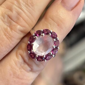 May include: A silver ring featuring a large, oval-cut, pale pink gemstone at the center, surrounded by a halo of twelve smaller, round-cut, deep magenta-colored gemstones set in a metal band.