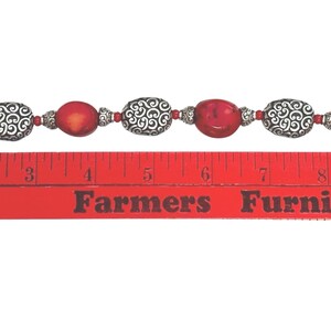May include: A bracelet featuring red oval beads and silver-colored, patterned beads. The bracelet is approximately 8 inches long, with a silver-colored chain and clasp. The beads are strung together with small silver-colored beads.