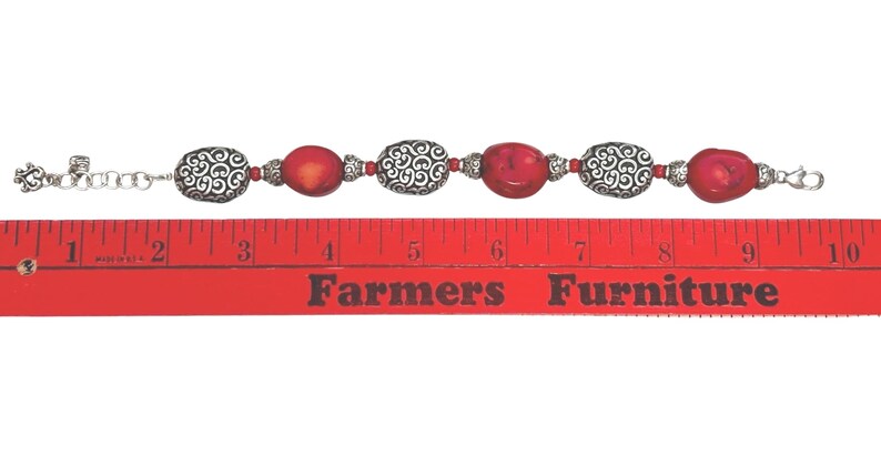 May include: A bracelet featuring red oval beads and silver-colored, patterned beads. The bracelet is approximately 8 inches long, with a silver-colored chain and clasp. The beads are strung together with small silver-colored beads.
