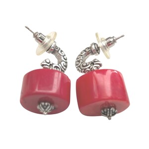 May include: A pair of earrings featuring a vibrant red, cylindrical bead. The earrings have a silver-toned, curved accent with an ornate design and a post back. The bottom of each bead has a silver-toned decorative cap.