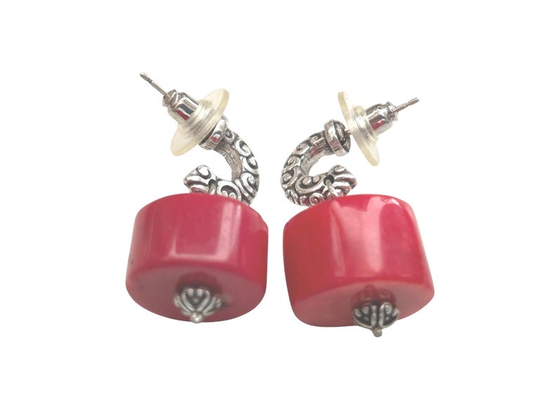 May include: A pair of earrings featuring a vibrant red, cylindrical bead. The earrings have a silver-toned, curved accent with an ornate design and a post back. The bottom of each bead has a silver-toned decorative cap.