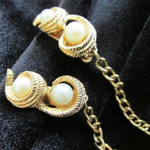 May include: A pair of gold tone, faux pearl,  swirl shaped,  brooches connected by a gold chain.