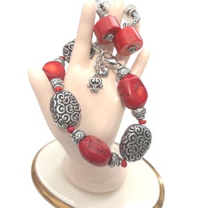 May include: A red and silver bracelet and matching earrings set. The bracelet features large red beads interspersed with silver filigree beads. The earrings have red barrel-shaped beads. The jewelry is displayed on a hand-shaped stand.