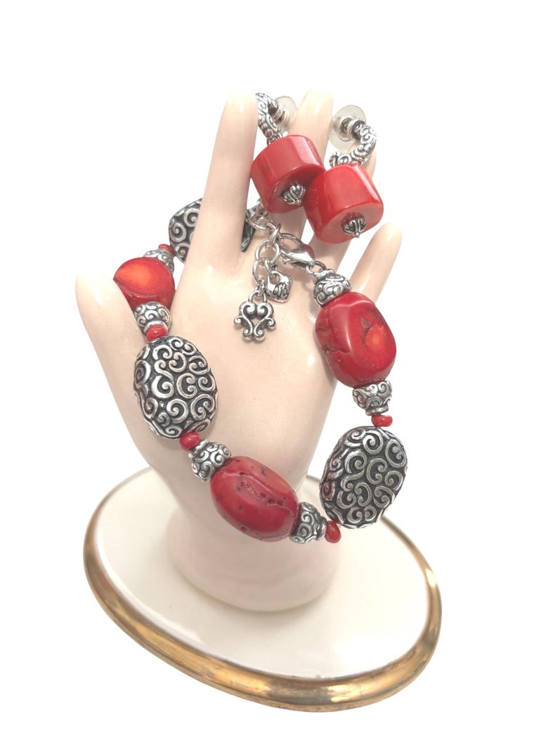 May include: A red and silver bracelet and matching earrings set. The bracelet features large red beads interspersed with silver filigree beads. The earrings have red barrel-shaped beads. The jewelry is displayed on a hand-shaped stand.