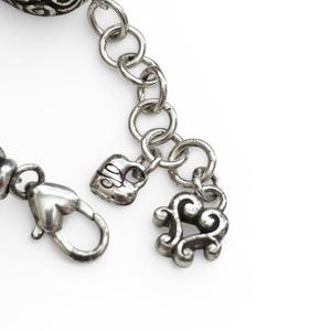 May include: Close-up of a silver-toned bracelet with intricate details. The bracelet features a heart-shaped clasp, a heart-shaped charm, and a decorative charm with a swirling design. The chain links are round and connected.