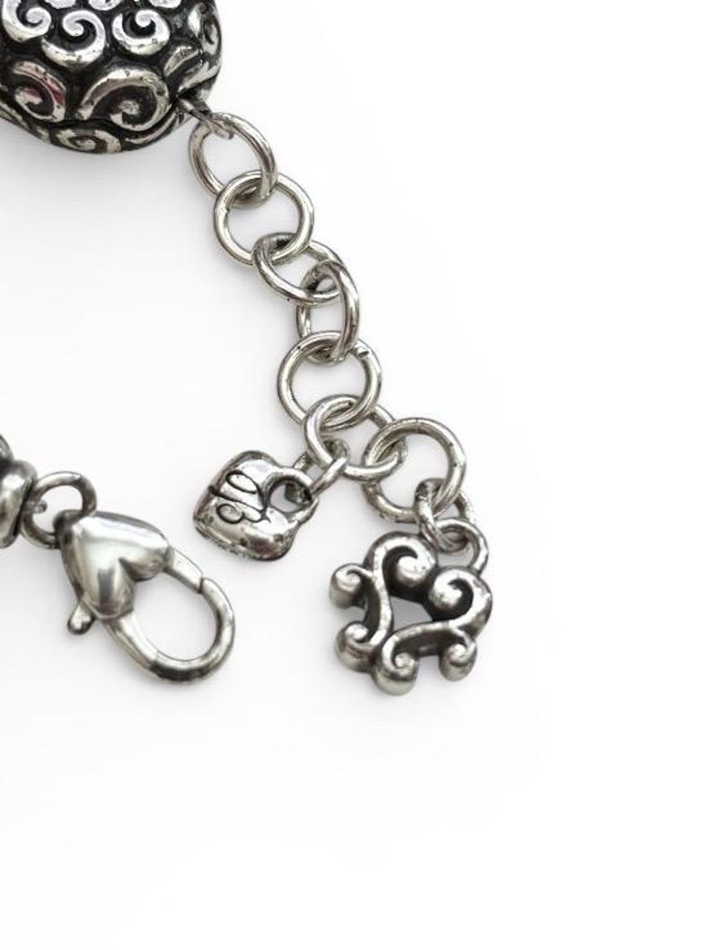 May include: Close-up of a silver-toned bracelet with intricate details. The bracelet features a heart-shaped clasp, a heart-shaped charm, and a decorative charm with a swirling design. The chain links are round and connected.