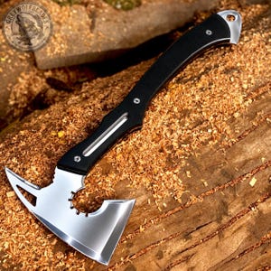 May include: A tactical hatchet featuring a polished silver metal blade with a cutout design and a gear-like detail near the neck. The handle is black with two silver rivets and a lanyard hole at the end. The logo Grey Wolf Blades is visible.