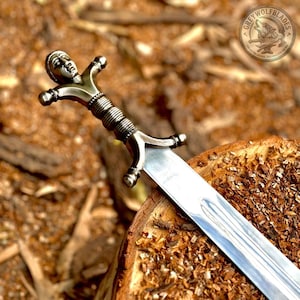 May include: A metal sword featuring a pommel shaped like a human head with detailed facial features. The hilt has a ribbed grip and a crossguard with rounded ends. The blade is silver-coloured, polished, and features a central fuller groove.