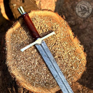 May include: A sword with a patterned Damascus steel blade, a polished brass crossguard, and a dark reddish-brown wooden handle with a brass pommel cap. The blade features a distinct wavy metal texture and a central fuller running down the length.
