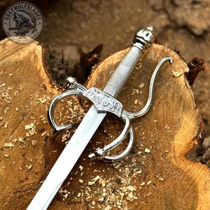 May include: A silver-toned rapier sword featuring a polished metal blade, a textured grip, and an ornate crossguard with curved quillons. The hilt displays intricate engravings, and the pommel is rounded. A circular logo reads GREY WOLF BLADES.