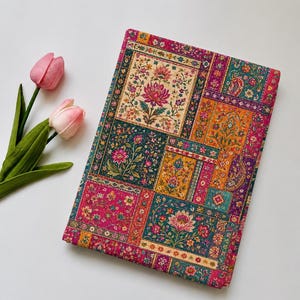 Puede incluir: Un cuaderno rectangular con un diseño de patchwork vibrante de patrones florales y de cachemira en tonos magenta, naranja, verde azulado y dorado. Dos tulipanes de color rosa suave con tallos y hojas verdes se sitúan a la izquierda.