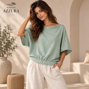 Op de afbeelding: Een saliegroene top met korte mouwen, een brede boothals en een geplooide elastische zoom, gecombineerd met een lichtbeige broek met wijde pijpen. De top heeft een losse, ontspannen pasvorm met verlaagde schouders en een textuur in de stof.