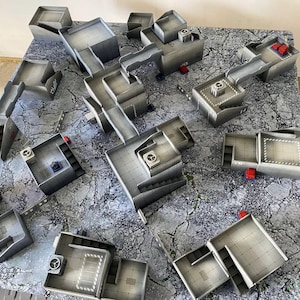 Peut inclure: Un ensemble de pièces de terrain modulaires en plastique gris pour jeux de plateau, comprenant diverses sections de bâtiments, des passerelles et des escaliers. Les structures ont une finition métallique avec des accents rouges et bleus.