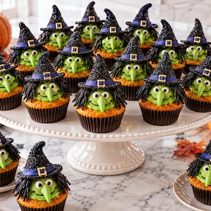 May include: A collection of cupcakes decorated as green-faced witches with black pointed hats, purple bands, and gold buckles. Each cupcake features green frosting, a pointed nose, and black hair, arranged on a white pedestal and small plates.