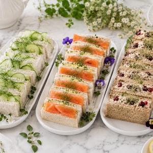 May include: Three white ceramic platters arranged on a marble surface, each holding a row of finger sandwiches. The left platter features cucumber sandwiches with dill, the center has smoked salmon, and the right displays chicken salad with cranberries.