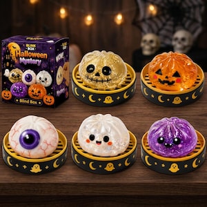 May include: A Halloween Mystery Blink Box set featuring five squishy dumpling-shaped toys in individual black and yellow steamer baskets. The toys include a gold glitter skull, an orange jack-o'-lantern, a bloodshot eyeball, a white ghost, and a purple creature.