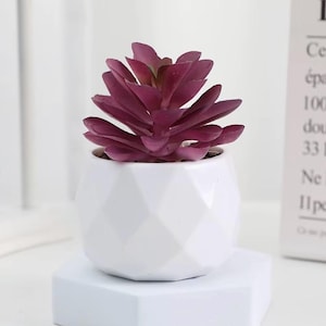 Puede incluir: Una pequeña planta suculenta artificial con hojas puntiagudas de color púrpura intenso dispuestas en forma de roseta. La planta está en una maceta de cerámica blanca geométrica con una superficie facetada de varios lados y acabado mate.