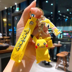 Puede incluir: Un llavero amarillo que presenta una figura 3D de Pikachu, una correa amarilla plana con el texto azul y amarillo Pokémon, y un dije blanco más pequeño con el texto amarillo Pikachu, todo unido a una argolla y cadena de metal plateado.