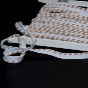 May include: A spool of white elastic trim featuring a repeating diagonal pattern of soft tan fuzzy stripes. The trim is wound around a white plastic card, with a loose end extending outward and forming a small loop on the surface.