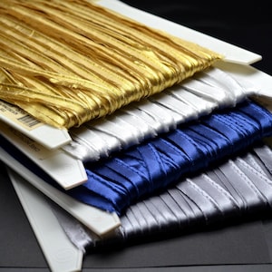 May include: Four spools of satin ribbon stacked vertically, featuring metallic gold, solid white, deep royal blue, and silver grey colours. Each ribbon is wound around a flat white plastic card, displaying a smooth, reflective texture.