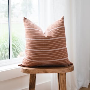 May include: A brown and white striped cushion sits on a wooden stool in front of a window with white curtains.