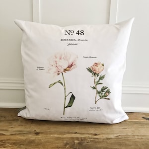 May include: White pillow with a botanical illustration of a pink peony flower. The pillow features the text "Nº 48 BOTANICA-Peonia -poksine- Pronia Mountes PLATE XIV piviese en arbre Bois. Coteaux Herbeux Fleurit d'avril en juin"