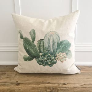 May include: Square throw pillow with a botanical design featuring various green cacti and succulents. The pillow has a natural linen-like texture and a neutral background, creating a rustic aesthetic. The cacti are detailed with shades of green and white.